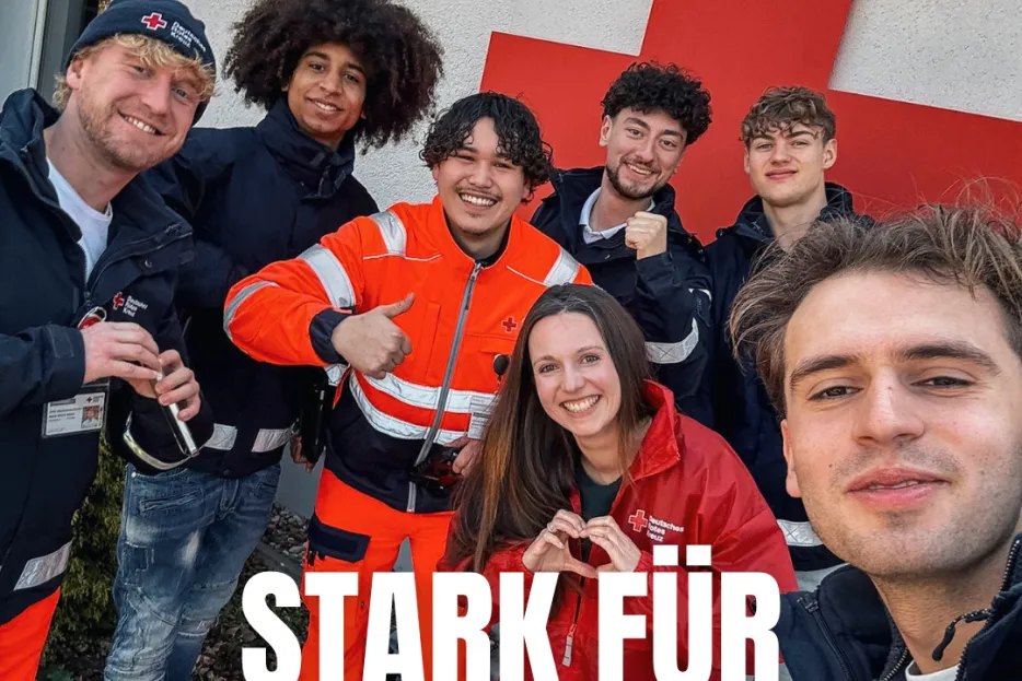 7 Personen zu sehen, davon 6 Männer und eine Frau mit DRK Begleitung, lächelnd in Kamera